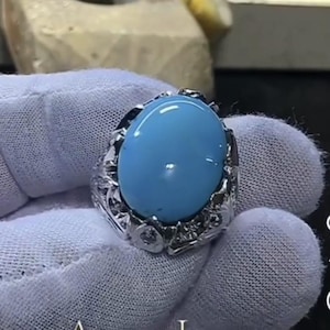 May include: A silver ring featuring a large, oval-shaped, light blue gemstone. The ring has an ornate, textured setting with small, clear accent stones. The gemstone is polished and has a smooth, glossy surface.