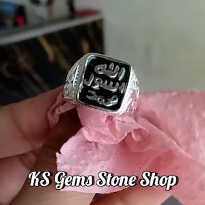 May include: A silver ring with a black square center featuring Arabic calligraphy. The ring has ornate detailing on the band and is held against a pink cloth. The text "KS Gems Stone Shop" is at the bottom.