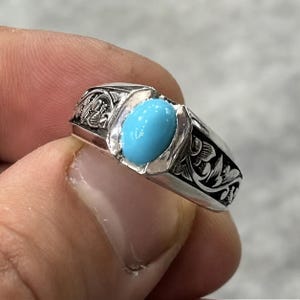 Op de afbeelding: Een zilveren ring met een centrale, ovaalvormige, felblauwe turkoois steen. De ringband is voorzien van complexe, donker geoxideerde florale gravures die langs de zijkanten doorlopen, wat zorgt voor een contrastrijk decoratief patroon op het gepolijste zilver.