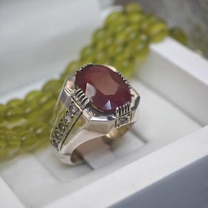 Op de afbeelding: Een zilveren ring met een grote, ovaal geslepen robijn. De ring heeft kleine accentstenen en ingewikkelde details op de band. De robijn heeft een dieprode kleur en de ring wordt gepresenteerd in een witte doos.