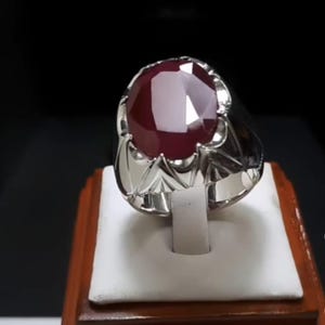 Puede incluir: Un anillo de plata con una gran gema de rubí ovalada y facetada. El rubí es de un color rojo intenso y rico. El anillo tiene un engaste de plata detallado y ornamentado con un diseño decorativo alrededor de la gema.
