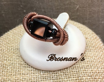 May include: A copper wire wrapped ring with a black square gemstone and black beads. The ring is displayed on a white ring stand.