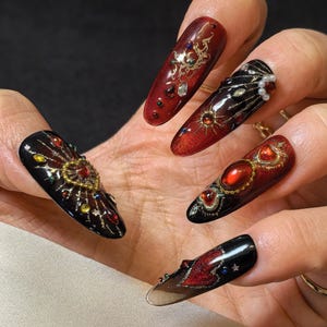 Può includere: Primo piano di cinque unghie finte lunghe a forma di mandorla. Ogni unghia presenta un design sfumato rosso scuro e nero, adornato con intricati filigrane dorate, strass e gioielli a forma di cuore. Le unghie sono una sorprendente esposizione di nail art.