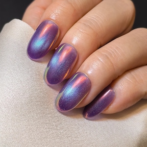 Peut inclure: Gros plan sur une main avec des ongles vernis. Les ongles sont de forme ovale et peints avec un vernis violet chatoyant et irisé qui reflète des teintes de bleu et de rose. La main repose sur un tissu de satin blanc.