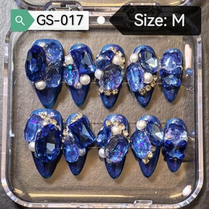 May include: A set of ten artificial nails in a clear plastic case. The nails are a deep blue with sparkling glitter and large, faceted blue gems, pearls, and gold accents. The text "GS-017" and "Size: M" are visible.