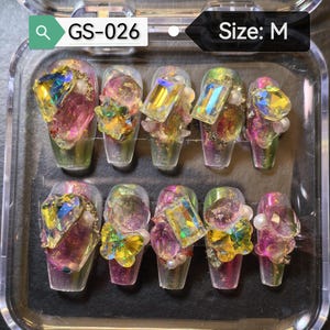 Puede incluir: Un juego de diez uñas artificiales en una caja de plástico transparente. Cada uña está adornada con pedrería iridiscente, perlas y gemas coloridas. Las uñas tienen un efecto degradado, pasando del verde al morado. El texto "GS-026" y "Size: M" son visibles.