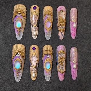 Puede incluir: Una colección de diez uñas artificiales largas y ornamentadas. Cada uña presenta un adorno dorado en la base, con diseños intrincados, gemas y una variedad de colores, incluyendo morado, rosa y blanco. Algunas uñas tienen un efecto nacarado.