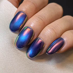 Puede incluir: Primer plano de uñas manicuradas con un acabado iridiscente. Las uñas de forma ovalada muestran una mezcla de azul, morado y toques de naranja y verde, creando un efecto de cambio de color. Las uñas se colocan sobre una tela suave de color blanquecino.