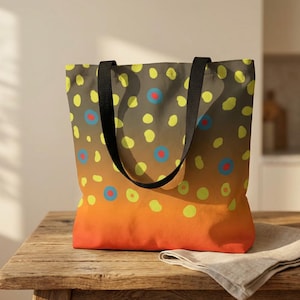 May include: A tote bag with a brown, orange, and yellow gradient design. The bag features a pattern of yellow and blue dots with red centers, and black handles. The bag is sitting on a wooden table.