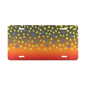 May include: Rectangular license plate with a gradient of orange to brown, speckled with yellow and blue-ringed dots. The plate has four slots for mounting and is designed with a fish-scale pattern.