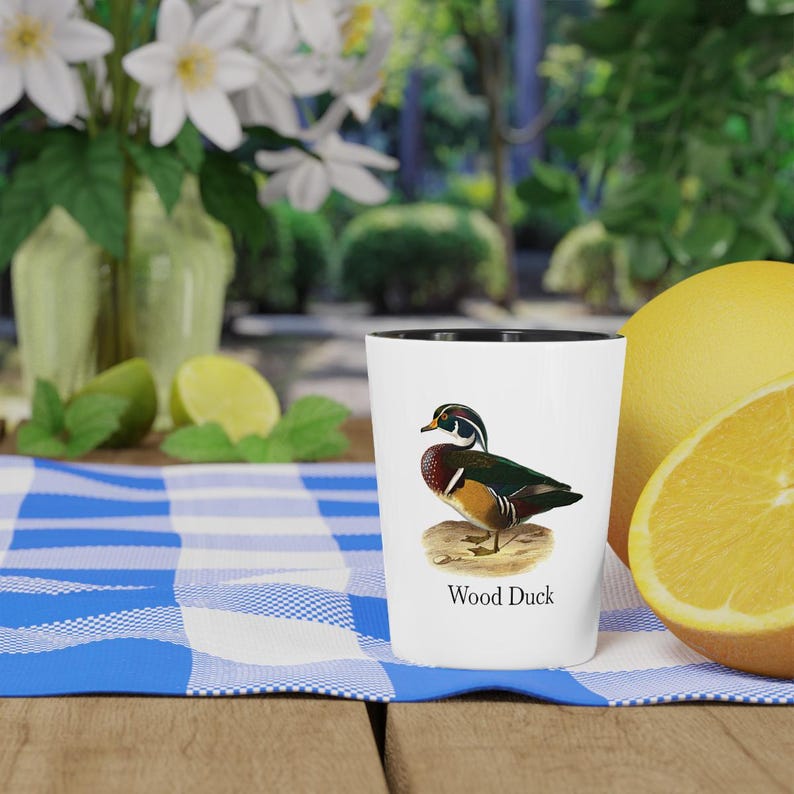 Puede incluir: Un vaso de chupito blanco con borde negro, con una ilustraci&oacute;n de un pato joyuyo. El pato tiene plumas verdes, marrones y rojas. Las palabras "Wood Duck" est&aacute;n impresas debajo de la ilustraci&oacute;n.