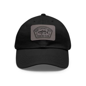 May include: Black baseball cap with a curved brim. A gray rectangular patch on the front features the text "AINT CAUGHT SHIT FISHING CLUB" with a fish graphic and two hooks. The cap is made of a soft, durable material.