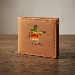 May include: A brown leather wallet featuring a colorful illustration of a fisherman casting a line. The image includes the text "On the Rise" below the illustration. The wallet is closed and appears to be made of a textured leather material.