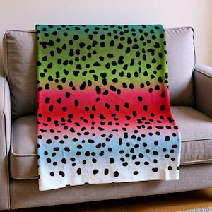 May include: A colorful blanket with a gradient design of green, red, blue, and white, adorned with black polka dots. The blanket is draped over a gray sofa, showcasing its vibrant pattern and soft texture. This blanket is perfect for adding a pop of color to any living space.
