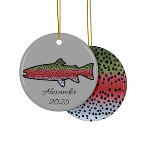 May include: A round, gray ornament with a colorful fish illustration. The fish is red, green, and black. The name "Alexander" and the year "2025" are printed below the fish. The ornament hangs from a gold string.