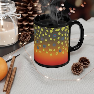 May include: A black ceramic mug with a colorful pattern of yellow, blue, and red dots over a gradient of brown to orange. The mug is filled with a dark liquid, and steam rises from it. The mug is on a light gray surface with decorative items.