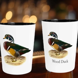 Puede incluir: Dos vasos de chupito blancos con borde negro presentan una ilustraci&oacute;n detallada de un pato joyuyo. El pato tiene un plumaje colorido, que incluye verde, rojo y amarillo. Un vaso tiene el texto "Wood Duck" debajo.
