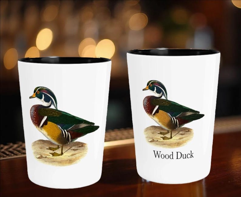 Puede incluir: Dos vasos de chupito blancos con borde negro presentan una ilustraci&oacute;n detallada de un pato joyuyo. El pato tiene un plumaje colorido, que incluye verde, rojo y amarillo. Un vaso tiene el texto "Wood Duck" debajo.