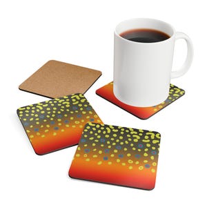 May include: A white ceramic mug filled with dark liquid sits on a coaster with a brown trout pattern. The coaster has a gradient of orange to yellow with dark spots and blue and yellow circles. Additional coasters are visible.