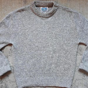 May include: A heather gray sweater with a crew neck and long sleeves. The sweater has a textured knit and a small label on the collar. The garment is laid flat on a patterned surface. A classic, casual style.