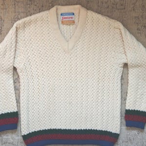 May include: Cream-colored V-neck sweater with a textured knit pattern. The cuffs and hem feature stripes in green, burgundy, and blue. The label reads "Jantzen". A classic, warm sweater.