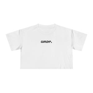 以下が含まれることがあります： 白いクロップトップ、半袖。 「amor」の文字が黒い筆記体でシャツの前面にプリントされています。 シャツはクルーネックで、裾はストレートです。