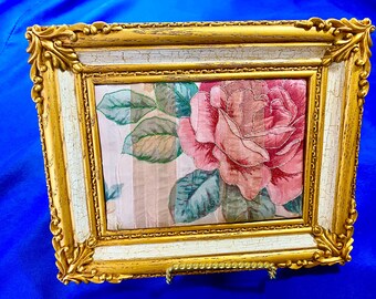 Op de afbeelding: Een ingelijst bloemenkunstwerk met een grote roze roos met groene bladeren tegen een gestreepte achtergrond. De lijst is sierlijk, met een gouden afwerking en een witte binnenrand met een verweerde uitstraling. Het kunstwerk wordt op een blauw oppervlak getoond.