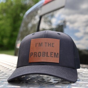Op de afbeelding: Zwarte baseballcap met een bruin leren patch met de tekst "I'M THE PROBLEM". De cap ligt op een metalen oppervlak, met een vage achtergrond van een voertuig en bomen.