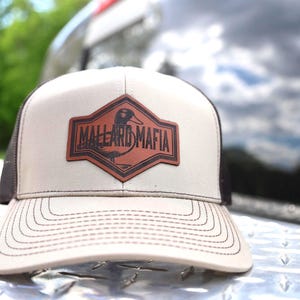 Puede incluir: Gorra de camionero beige y negra con un parche de cuero marrón. El parche presenta una ilustración de un pato y las palabras "MALLARD MAFIA". La gorra tiene una visera curva y una parte trasera de malla.