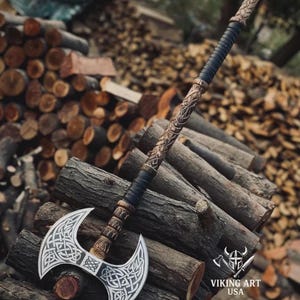 May include: A double-bladed axe with a silver blade and intricate Celtic knotwork design. The handle is wrapped in black leather and features carved wooden accents. The axe rests on a pile of wooden logs, with the "Viking Art USA" logo visible.