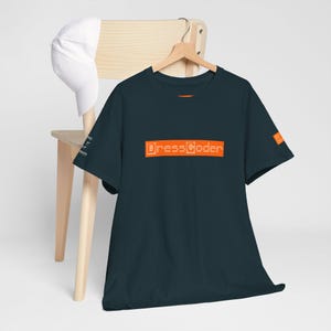 May include: A dark teal t-shirt with the text "DressCoder" in orange block letters. The shirt is hanging on a wooden hanger. A white baseball cap rests on a wooden chair behind the shirt.