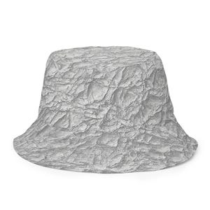 Puede incluir: Un sombrero de pescador con un patrón de roca texturizado en gris claro en toda la superficie. El sombrero tiene una parte superior plana, una corona cilíndrica y un ala inclinada hacia abajo, con un estampado realista de superficies rocosas.