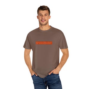 Peut inclure: Un t-shirt marron avec le texte "DressCoder" en lettres orange. Le t-shirt est fait d'une matière douce et est porté par une personne. La personne porte un jean bleu.