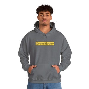 Op de afbeelding: Een grijze hoodie met een geel rechthoekig logo met de tekst "DressCoder". De hoodie heeft een voorzak en een klein logo op de linkermouw. De persoon draagt een blauwe jeans.