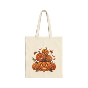 May include: A beige canvas tote bag featuring a central graphic illustration of a stack of five orange jack-o'-lantern pumpkins with carved faces, accented by small brown autumn leaves and tiny flying bats scattered around the pumpkin cluster.