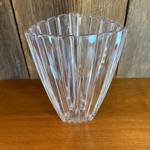 May include: A clear, faceted glass vase with a flared, square shape. The vase has vertical ridges that create a textured surface. The vase is sitting on a wooden surface.