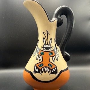 May include: A hand-painted ceramic pitcher with a cream-colored body, a black handle, and a reddish-orange base. The pitcher features a Native American-inspired design with a central figure in orange, black, and turquoise. The pitcher is approximately 7 inches tall.
