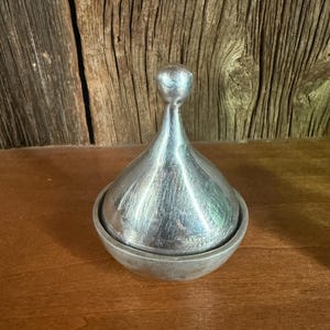 May include: A silver-colored, lidded container with a rounded base and a conical top. The lid has a small, spherical handle. The container sits on a wooden surface, with a wooden background.