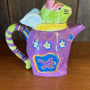 May include: A whimsical, hand-painted ceramic teapot featuring a cat-like creature with angel wings. The teapot is primarily purple with blue, yellow, green, and pink accents. The handle is striped, and the body is decorated with floral designs.