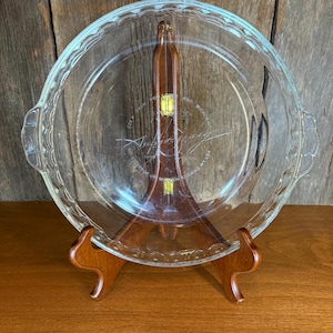 May include: A clear glass pie dish with a scalloped edge and handles, resting on a wooden stand. The dish has text that is partially visible. The stand is made of dark brown wood. The background is a wooden surface.