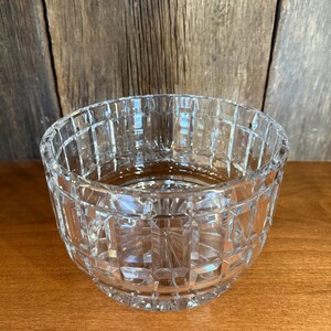 May include: A clear, cut-glass bowl with a geometric pattern. The bowl is round with a square-cut design and sits on a wooden surface. The background is a wooden wall.