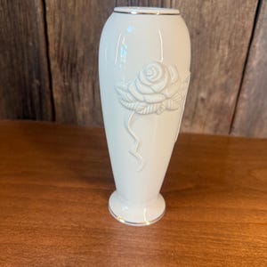 May include: A white ceramic vase with a raised rose design on the front. The vase has a gold trim around the top and base. The vase is approximately 8 inches tall.