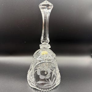 May include: A clear crystal bell with a long handle and a diamond-shaped top. The bell features a floral design etched on the front and a textured pattern throughout. The bell is a decorative item.