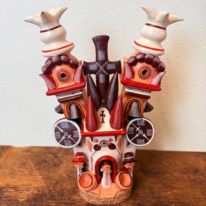 May include: A decorative clay sculpture featuring a central cross, two tall towers with white and red accents, and circular side elements. The base displays three small figures standing in arched openings, with painted geometric patterns in red, brown, and white.