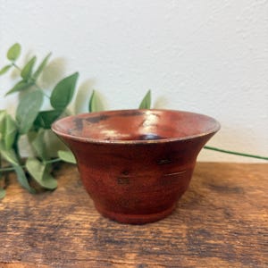 May include: A small, wide-mouthed ceramic bowl featuring a deep, mottled red glaze with dark brown and black undertones. The bowl has a flared rim, a rounded body, and a smooth, glossy finish that highlights the variations in the clay texture.