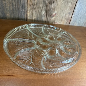 May include: A clear glass serving platter with a leaf design. The platter is round with a central, raised section and several divided compartments. The edges have a scalloped design. The platter is on a wooden surface.