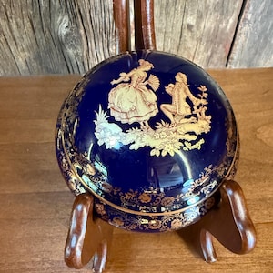 May include: A round, cobalt blue Limoges porcelain box with a gold floral design and a gold illustration of a couple in period clothing. The box is displayed on a wooden stand.