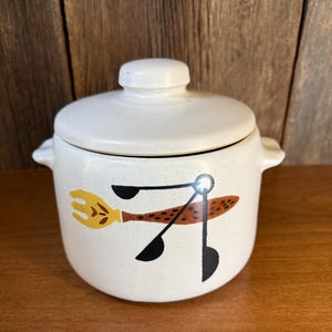 May include: Cream-colored ceramic canister with a lid and side handles. The canister features a speckled finish and a graphic of a yellow fork, brown spoon, and black tongs. A vintage kitchenware item.