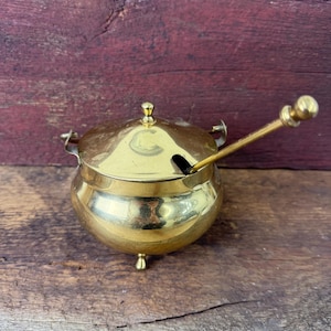 May include: A polished brass condiment pot with a rounded body, three small feet, and a matching lid. The pot features a small handle on each side and includes a long, slender brass spoon with a spherical finial resting in a side notch.