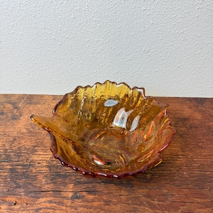 May include: An amber-colored glass bowl shaped like a leaf with a textured, crackled surface. The bowl features a scalloped edge and a small, clear sticker near the center that reads "LEKKO HANDCRAFT" in white text against a blue background.